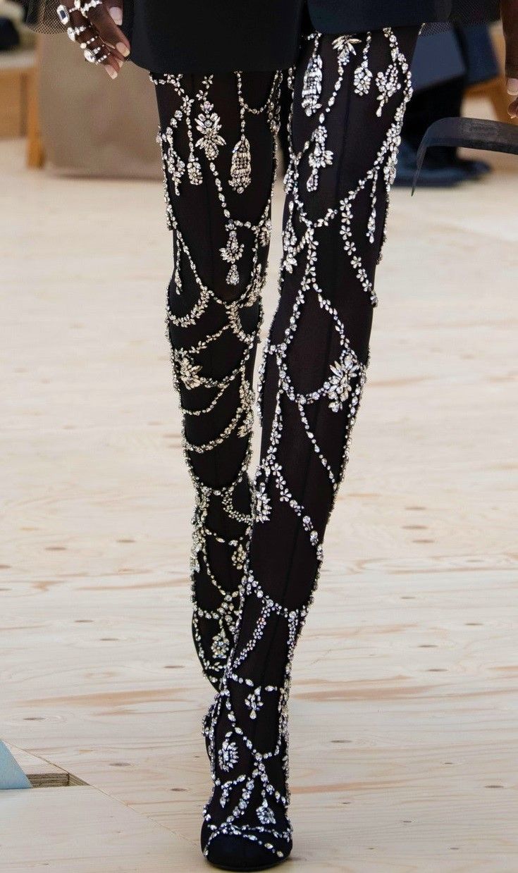 a closeup shot of a model's legs showcasing intricately embellished black tights and matching highheeled boots. the tights and boots are adorned with a dazzling array of crystals and rhinestones, creating a striking, luxurious effect. the overall style is elegant and glamorous, typical of highfashion runway presentations.  the model's hand, wearing multiple rings, is partially visible in the upper left corner. the background is blurred, focusing attention on the detailed embellishments.