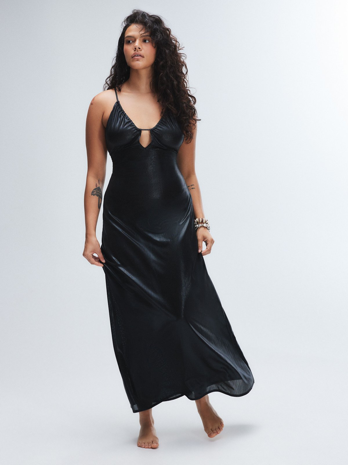 a fulllength shot of a woman model, against a lightgrey background. she is wearing a black, shiny, maxi slip dress with thin straps and a keyhole neckline.  her dark, curly hair is loose around her shoulders. she is barefoot and is wearing simple bracelets.  the overall style is elegant and simple.