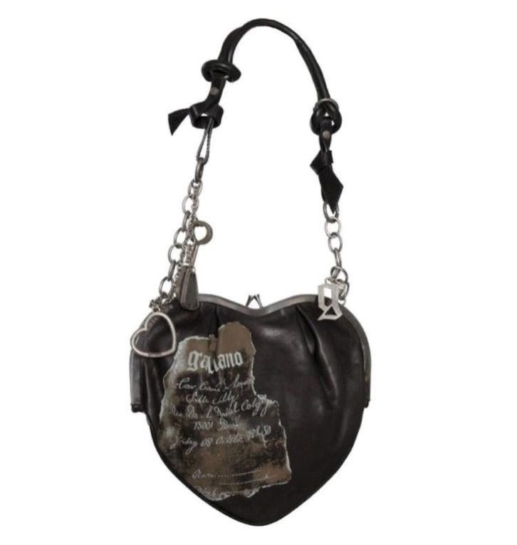 a heartshaped black handbag is suspended in the center of the frame. the bag features a metal frame at the top, along with a chain strap. the strap is adorned with multiple silver charms. a printed design is visible on the bag. the bag appears to be a john galliano design.