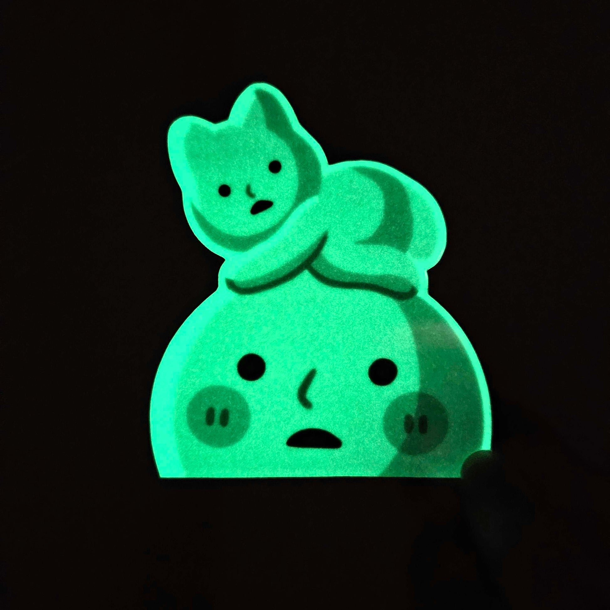 the image shows a glowinthedark sticker or decal. the sticker depicts a cartoonish moonlike character with a smaller cat sitting atop it.  both the moon and the cat are bright green, suggesting a phosphorescent material. the background is completely black, highlighting the glow of the sticker. the style is cute and whimsical.