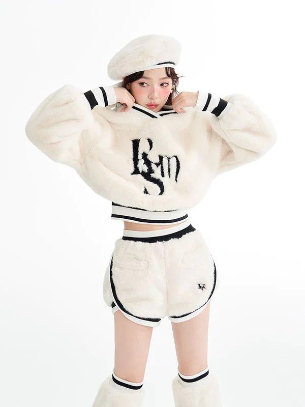 a young woman in a white fur sweatshirt and shorts with black trim poses in a studio. she is wearing a white beret and has her hair pulled back. the background is white.