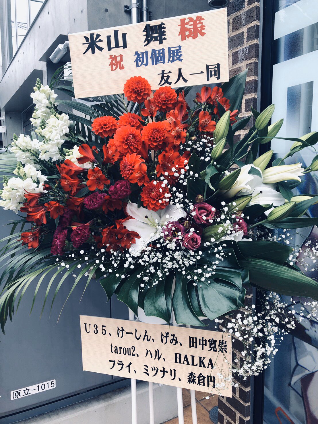 an image of a floral arrangement in celebration of someone's first solo exhibition. there is a sign that reads "congratulations on your first solo exhibition, mai yoneyama". the arrangement is made up of red, pink, and white flowers. there is also a smaller sign with a list of names.