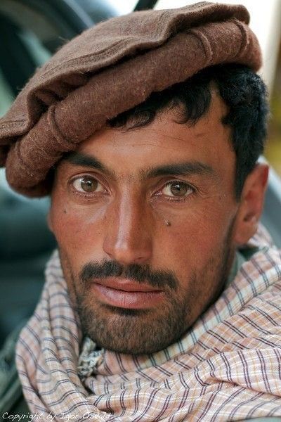 a portrait of a man wearing a pakol hat and a headscarf. the man has dark hair and brown eyes, and he is looking directly at the camera with a serious expression.