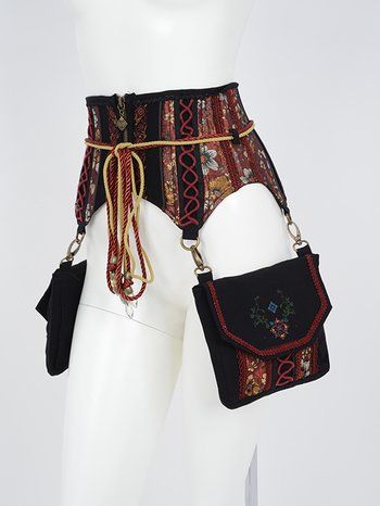 a waist/garter belt combination with attached pouches is displayed on a mannequin. the belt itself is a wide band, mostly black and decorated with a red floral pattern. it is secured with a zipper in the back, and a red and gold rope is tied around the belt, with tassels hanging down. there are two black pouches attached to the belt with metal clasps and chains. one pouch is a simple envelope style, while the other has a decorative floral embroidery and red trim.