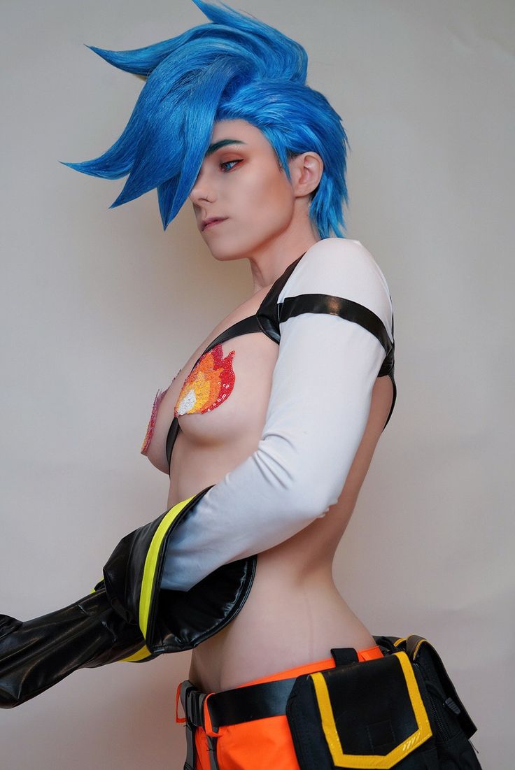 a person in a character cosplay costume with bright blue hair. they are wearing an outfit with orange, black, and white elements, and flame details on their chest.