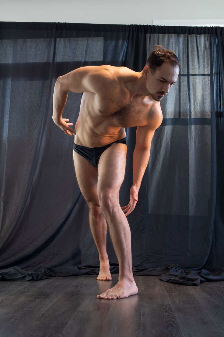 a muscular man in black briefs stands in a studio, facing towards the left. he has his left hand on his hip, and his right hand is down by his side, seemingly reaching for something. he is looking down at the ground with a slight frown, as if he is thinking hard. the background is dark black curtains, and the lighting is focused on the subject, creating dramatic shadows.