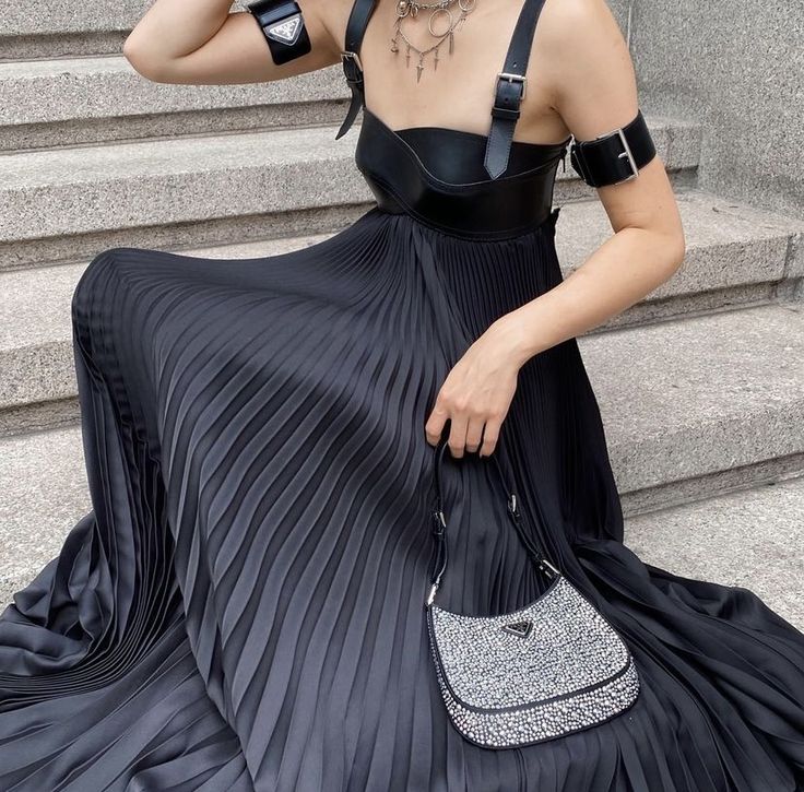 a woman is sitting on a set of stairs wearing a black dress with a leather bustier top and pleated skirt. she is accessorized with a layered necklace, an arm band, and a prada purse.