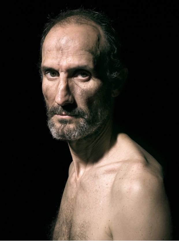 a dramatic portrait of a middleaged man with a gray beard, bare chest, and intense facial expression against a dark background. the image uses strong lighting to highlight the man's weathered skin and features.