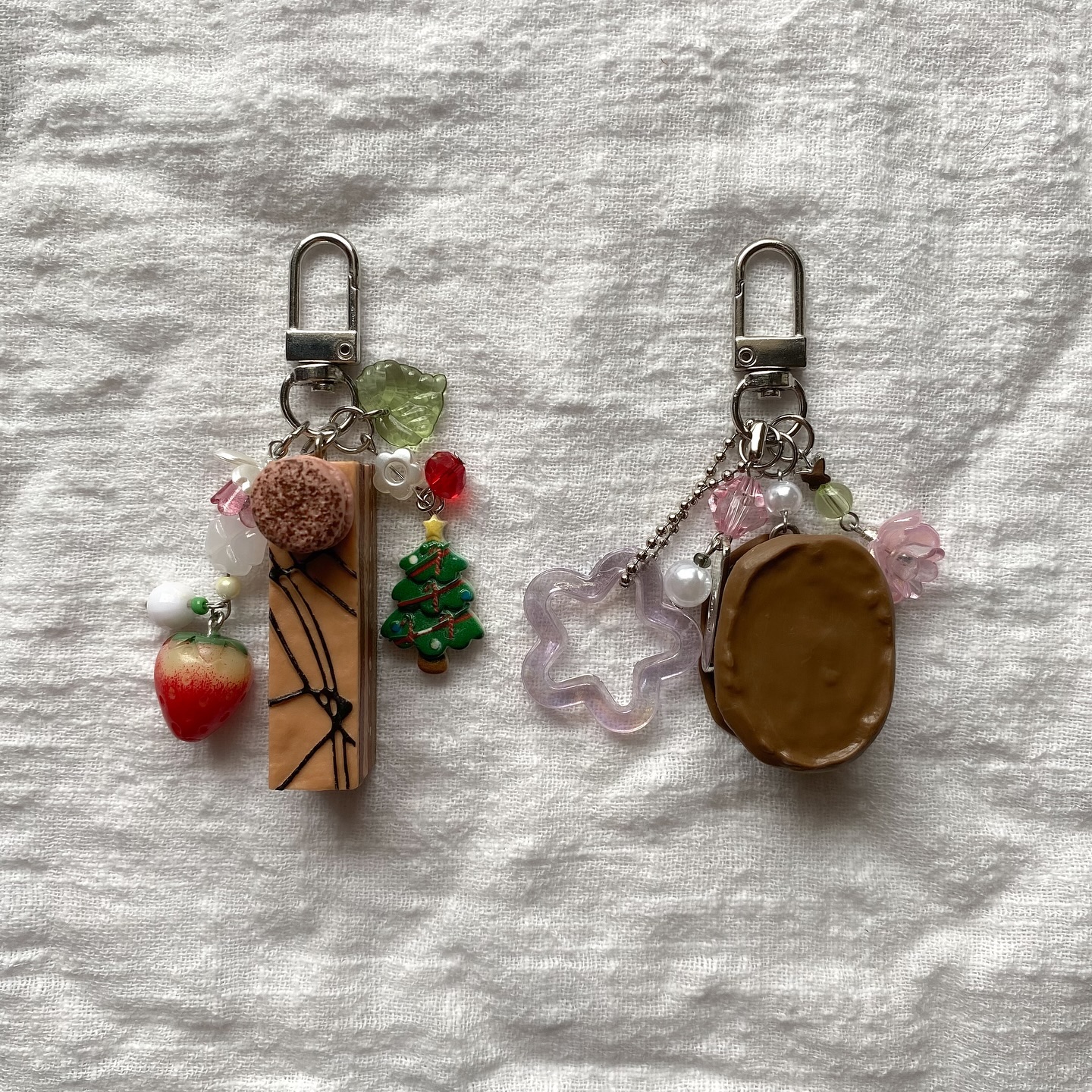 two charm keychains are laid on top of a white fabric surface. the keychain on the left features a charm designed to look like a rectangleshaped pastry with dark brown stripes, a brown ball charm, a strawberry charm, and a christmas tree charm with decorative beads. the keychain on the right has a charm that resembles a brown cookie or cake and a starshaped charm, also adorned with decorative beads. both keychains are attached to a standard keychain clip.