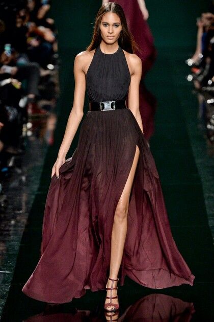 a fashion model walks the runway in a long black and red gown. the gown has a high neck and a belt around the waist. the model has long, straight hair and is wearing makeup. she is walking in heels.