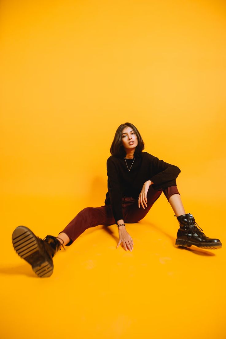 a young woman wearing black boots, black sweater, maroon pants is sitting on a yellow background. she is looking at the camera. her legs are stretched out in front of her, her left foot is pointing towards the camera.