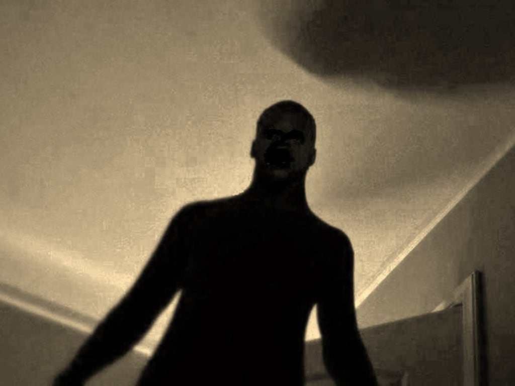 a silhouetted figure of a man is captured in a lowangle shot, creating a menacing and eerie atmosphere. the image is monochrome with a sepia tone, enhancing the sense of darkness and mystery. the man's features are obscured, adding to the unsettling nature of the scene, and the dramatic lighting further emphasizes the threatening mood. the setting appears to be indoors, with a ceiling and a door visible in the background.