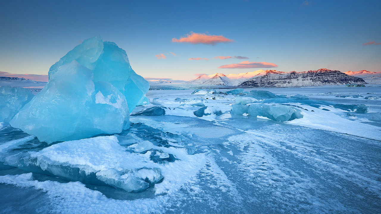 a panoramic view of a frozen lake with a large glacier in the foreground, surrounded by snowcovered mountains. the sky is a beautiful blue, and the clouds are a soft pink. the sunlight is reflecting off the ice and snow, creating a shimmering effect.  the scene is peaceful and serene, creating a sense of awe and wonder.