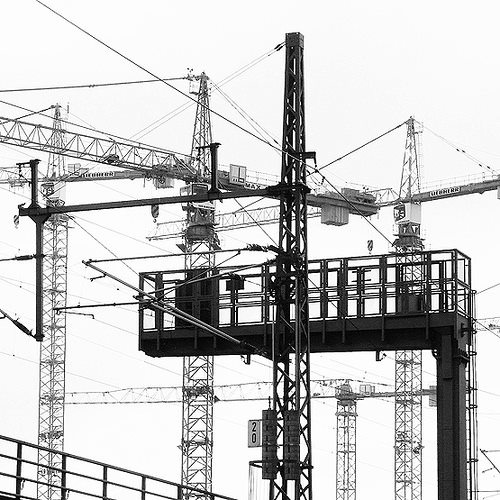 the black and white photograph depicts a complex interplay of several tall tower cranes against a backdrop of intricate overhead railway lines and electrical wires. the cranes, varying in height and design, suggest a largescale construction project underway in an urban setting.  a prominent metal gantry structure, possibly part of the railway infrastructure, occupies the foreground, partially obscuring some of the cranes. the image contrasts the organic, complex nature of the wire systems with the geometric precision of the manmade cranes and the sturdy railway gantry. the overall impression is one of intense industrial activity and urban development. the lack of color enhances the starkness of the metal structures and the web of lines.