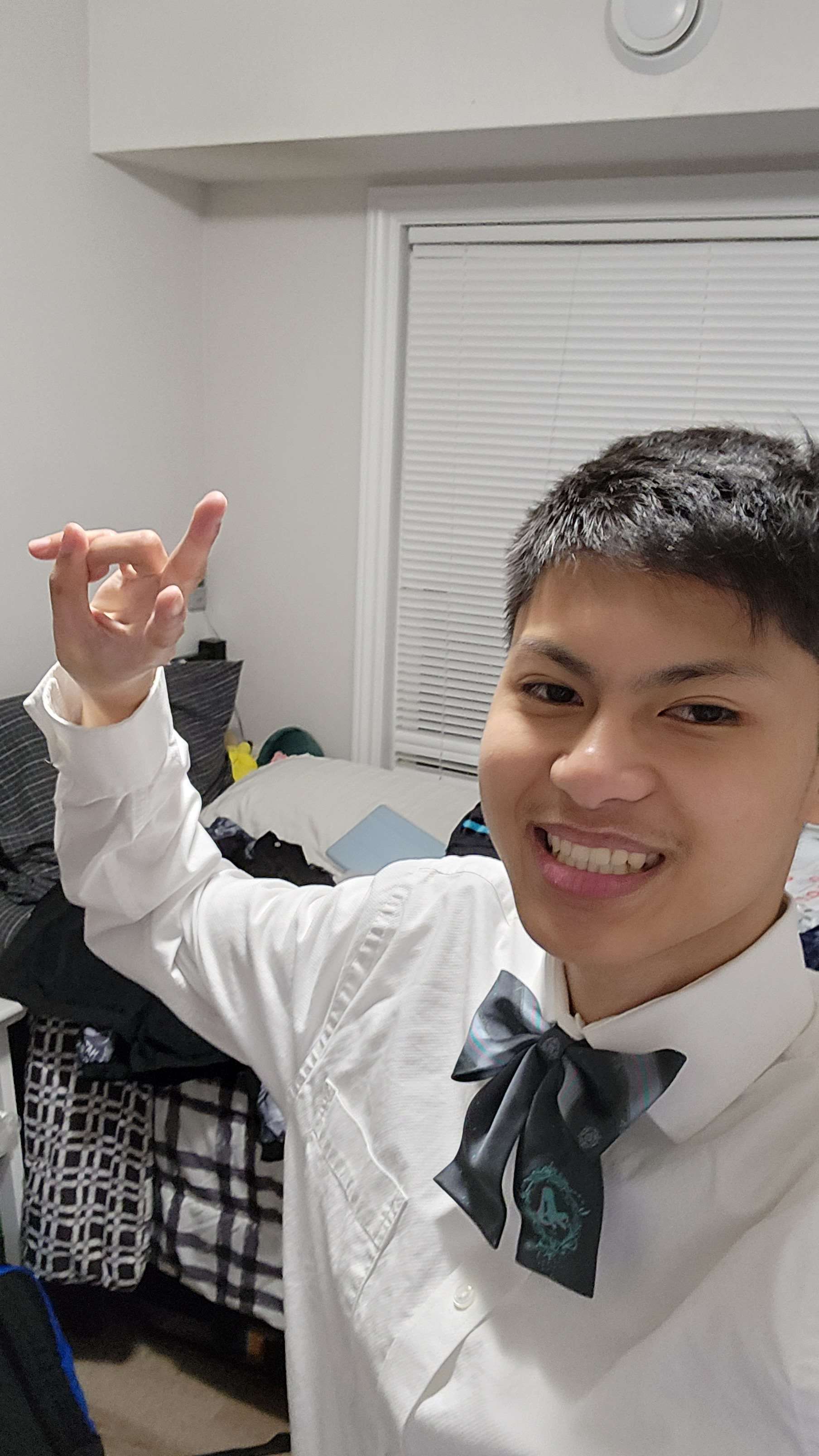 a young man is taking a selfie in his bedroom. he is wearing a white shirt and a bowtie. he is smiling at the camera.  the room is messy with clothes and bedding visible. the lighting is bright and natural coming from the window.