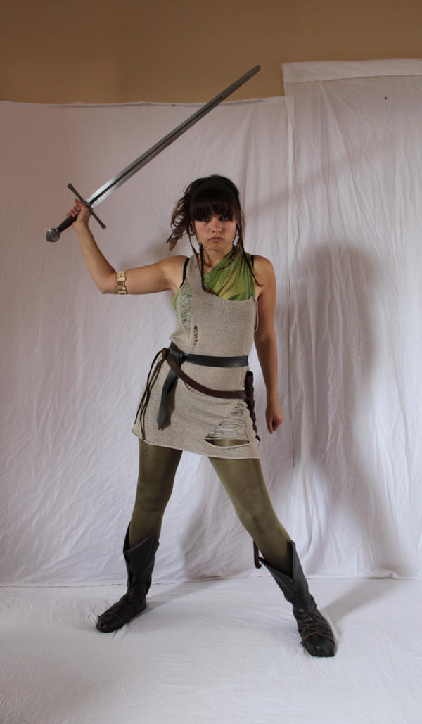a fullbody shot of a young woman dressed in a medievalstyle costume. she's holding a long sword in her right hand, her left hand is at her side.  the costume includes a ripped beige tunic, olive green tights, and dark brown boots. she has on a brown leather belt and a green scarf around her neck.  the background is a plain white sheet. the overall impression is that she is portraying a fantasy warrior or character.