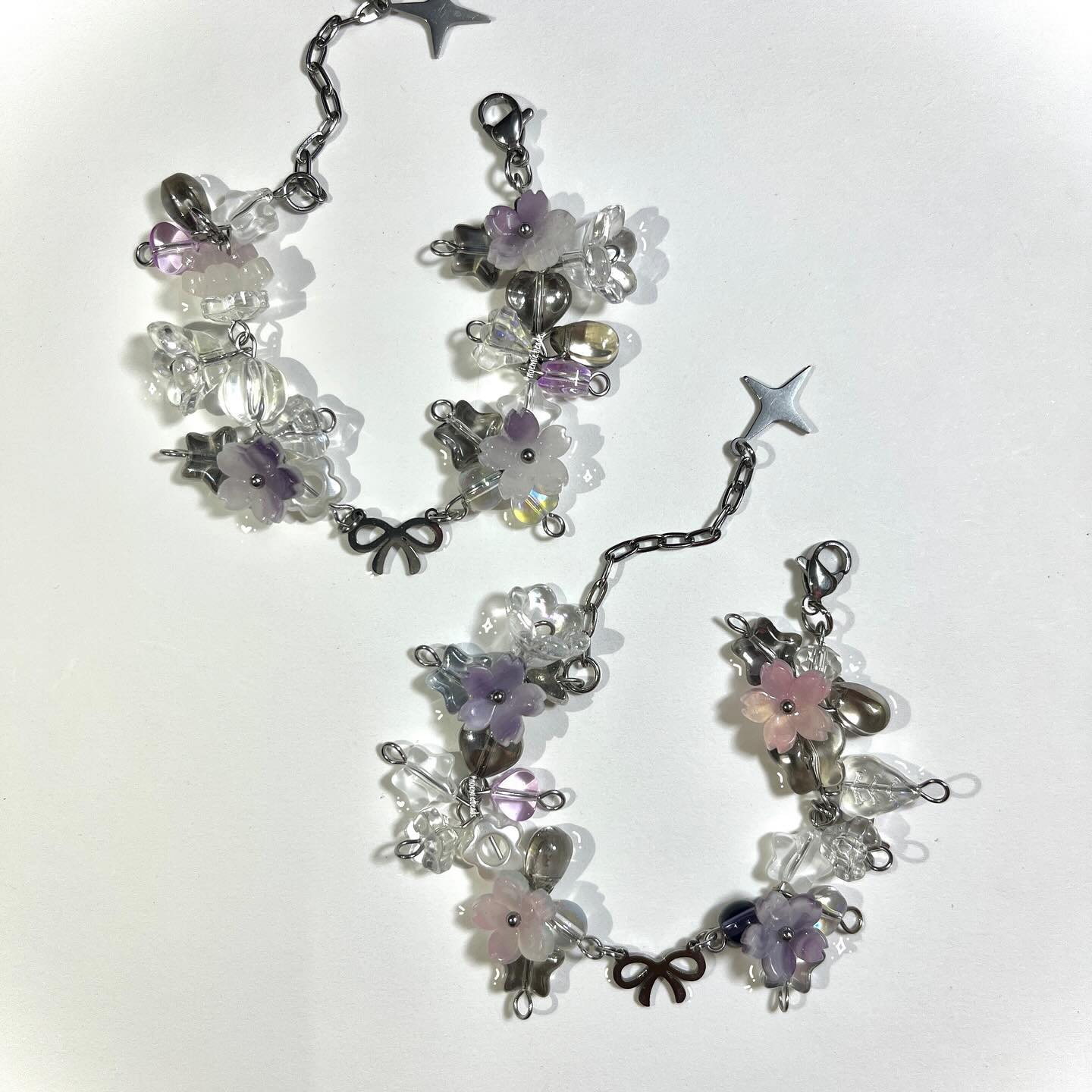 two handmade beaded bracelets with silver chains, flower beads, butterfly charms, star charms, and clasps are displayed on a white surface. the bracelets are made with a combination of clear, purple, and pink beads. they feature flowershaped beads, butterfly charms, star charms, and bow charms. the silver chains connect the beads and charms. the clasps are attached to one end of the chains.