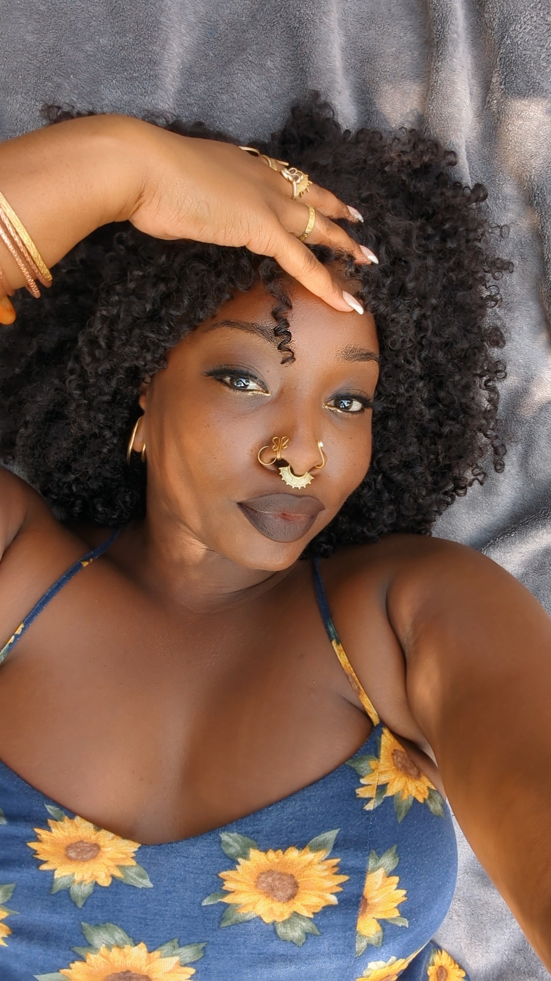 a young woman with dark skin and curly black hair is wearing a blue sundress with yellow sunflowers. she is smiling, looking at the camera, with her hand on her head. she has on gold jewelry, a nose ring, and earrings. she is sitting down and the background is a soft grey. she is looking happy and confident.