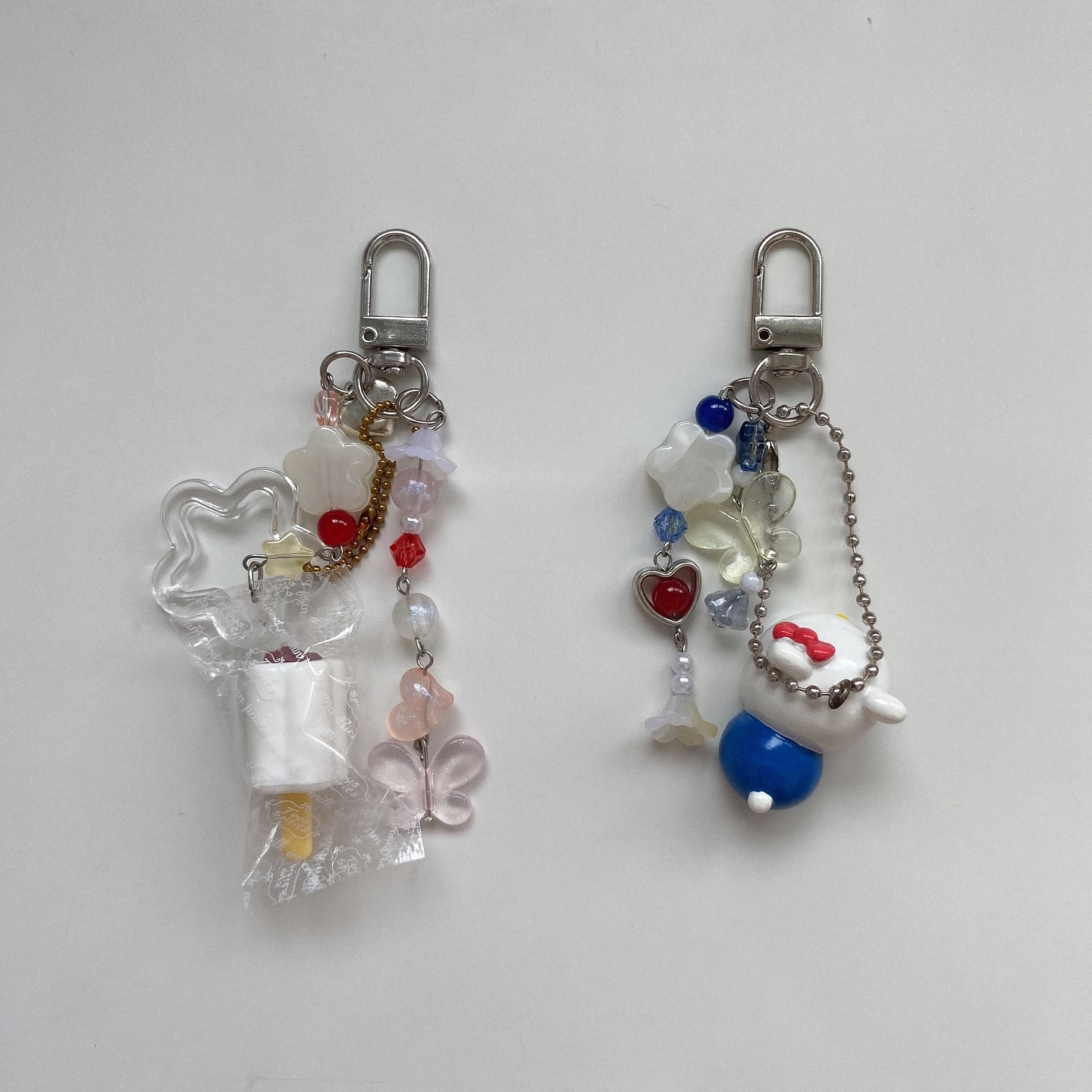 the image shows two keychains with various beads, charms, and silver hardware. the left keychain features a clear starshaped frame, a wrapped popsicle charm, and a string of pink, red, and gold beads, plus butterfly charms. the right keychain has a blue and white character charm, a chain, and a string of blue, white, and red beads and flower charms. both keychains have silver clasps.