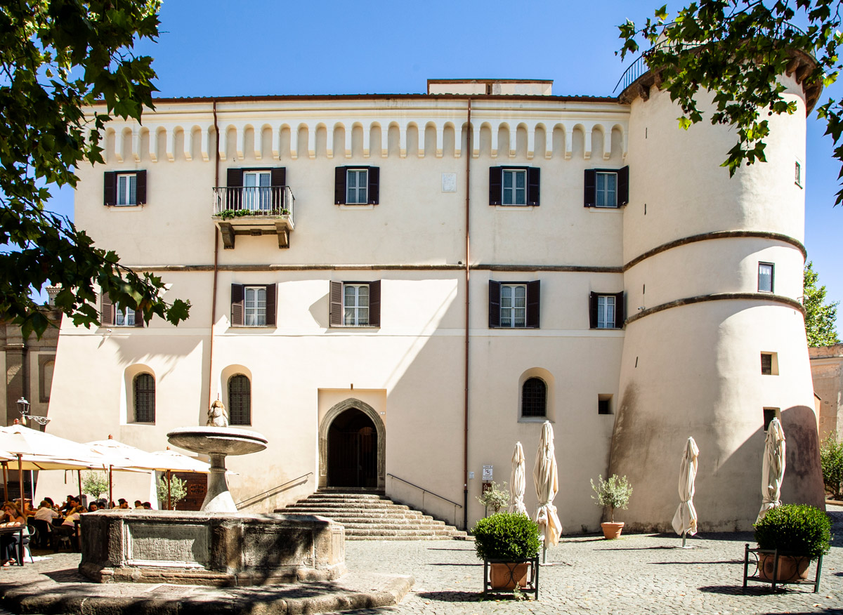 a medium shot of a medieval castle with a turret and a courtyard in front of it. there is a fountain in the courtyard, some closed umbrellas, and a restaurant area under open umbrellas. a small balcony is visible on the castle's second floor, and trees are visible on the left and right sides of the shot. the sky is blue.