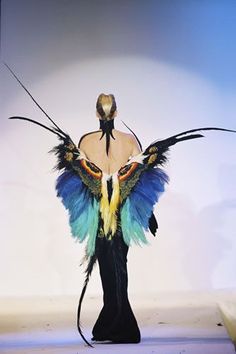 a woman model in a high fashion show with elaborate feathers in the colors of blue, green and black is standing on a runway, wearing a stylish black dress, with feathers forming a pair of wings that are flowing behind her. she has a serious expression on her face, her hair is styled and her make up is applied in a way that compliments her outfit.