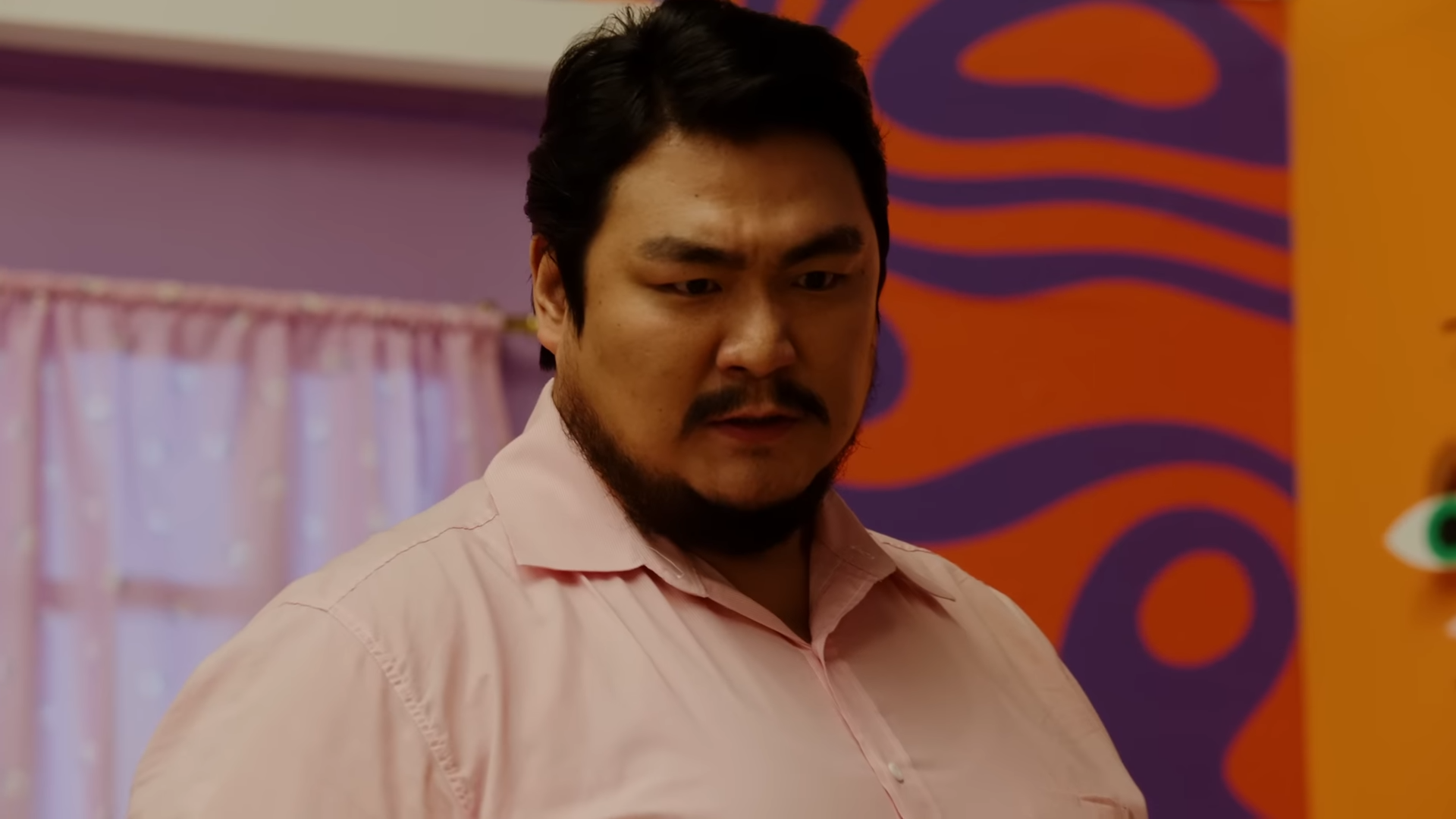 a closeup photograph of a man with a concerned expression. he has short black hair, a beard, and is wearing a pink collared shirt. the setting appears to be indoors with pink and orange colored walls.