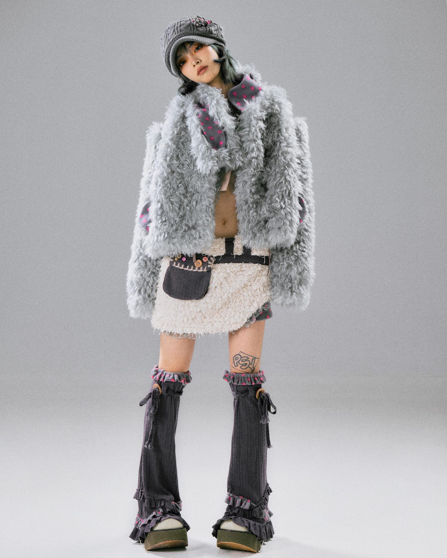 fullbody shot of a young woman modeling a fashionable outfit.  she's wearing a light grey fluffy coat, a creamcolored skirt with a brown pouch attached to the waist, dark grey leg warmers with ruffled detailing at the bottom and matching shoes, and a dark grey newsboystyle hat. the model has short, dark hair and subtle makeup. the background is a simple light grey, making the outfit stand out. the overall style suggests a blend of casual and somewhat whimsical elements.