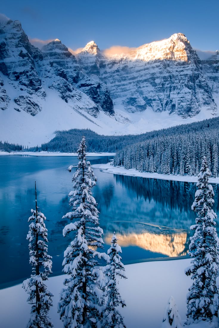 a scenic winter view of moraine lake in banff national park, alberta, canada. the lake is surrounded by snowcovered mountains and coniferous trees. the water reflects the mountains and the sky. snowcovered evergreen trees are in the foreground. the sky is a clear blue with some clouds. the tops of the mountains are illuminated by golden light.