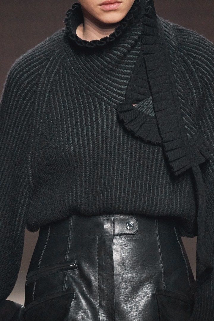 a woman is wearing a black sweater with a ruffle collar and a black highwaisted leather skirt. she is looking at the camera.