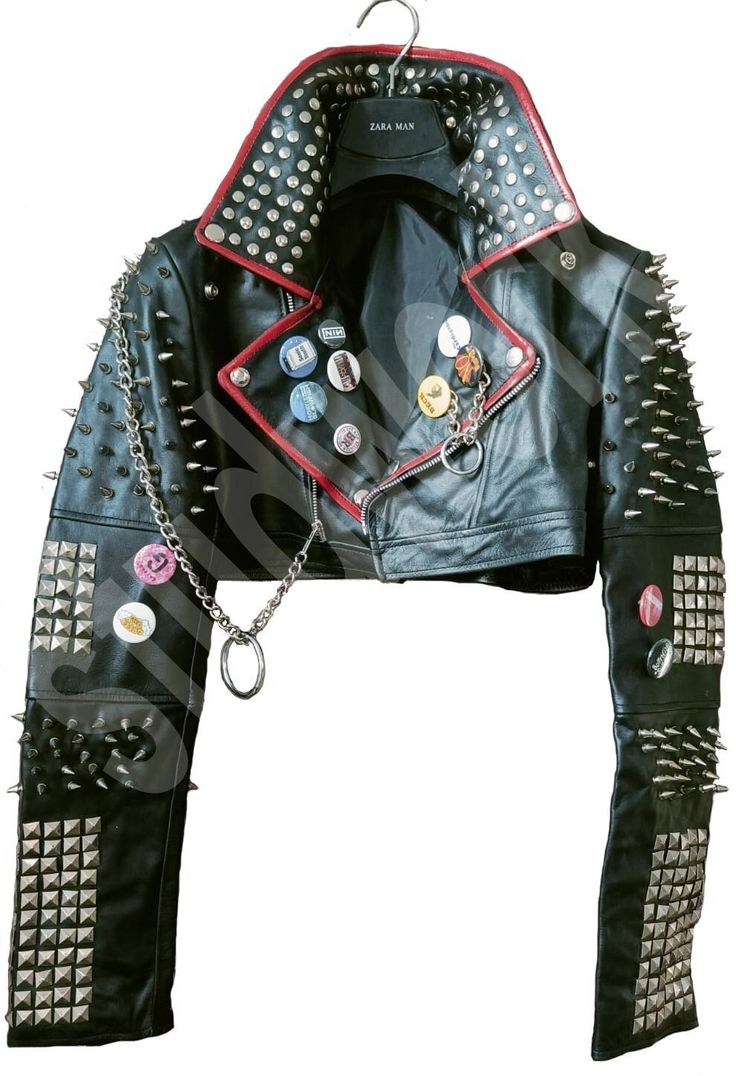 a black leather jacket with silver studs and spikes, red trim around the collar, and several pinback buttons on the left side. the jacket is hanging on a clothes hanger. the sleeves are covered in silver studs. the jacket has a silver chain hanging from the zipper.