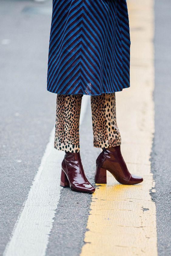 a closeup shot of a woman's legs, showcasing her stylish outfit. she is wearing a blue chevronpatterned coat, leopard print pants, and burgundy patent leather ankle boots with a block heel. the photo is taken on a city street, with a yellow line visible on the pavement. the overall style is chic and trendy.