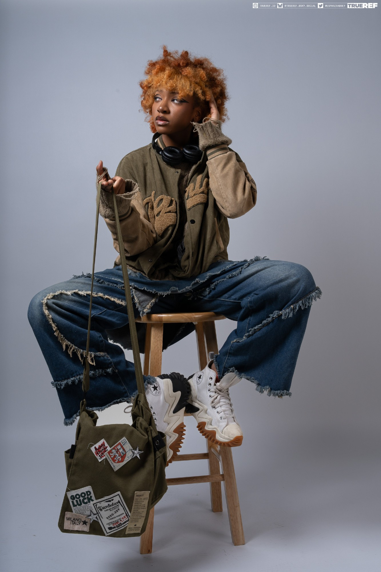 a young woman with vibrant orange curly hair is stylishly seated on a wooden stool against a muted grey backdrop. she's clad in a casual yet fashionable outfit, combining a distressed pair of denim jeans with an olive green bomber jacket. the jacket features unique patches and a relaxed, oversized fit. her footwear consists of striking white hightop platform converse sneakers. a stylish olive green canvas shoulder bag, also adorned with various patches, hangs from her shoulder, adding to the overall aesthetic.  headphones are draped around her neck, suggesting a contemporary, urban style. her pose is relaxed yet intentional, capturing a trendy and cool demeanor. the image is a professional studio shot with careful lighting and a neutral background that centers the focus on her attire and expression.