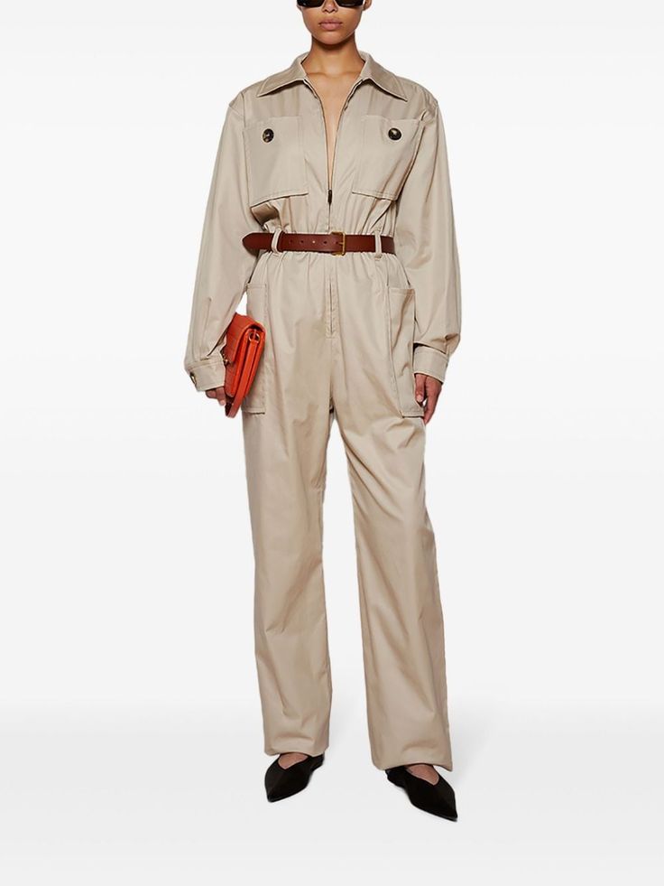 a woman is wearing a beige long sleeve jumpsuit with a zipper in the front. it has a belt around her waist and big pockets on the chest and legs. she is wearing black flat shoes and sunglasses. she is holding a small orange bag in her hand.