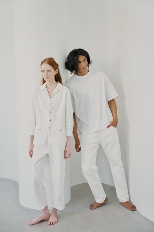 a full shot of a man and woman standing against a white wall. the woman is wearing a white denim jumpsuit and the man is wearing a white tshirt and white pants. both are barefoot. the image is minimalist and clean.