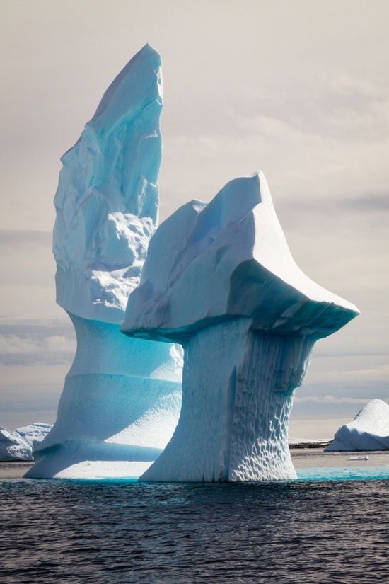 the image shows a large iceberg in the ocean. the iceberg is tall and has a unique shape, with a tall, thin spirelike structure on the left and a wider, mushroomshaped structure on the right. the ice is blue and white, and the water is dark. the background is a hazy sky. other smaller icebergs are also visible in the background.