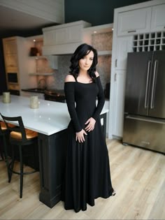 a woman in a black offtheshoulder dress with long sleeves stands in a modern kitchen. she is smiling and looking at the camera. the kitchen has white cabinets, a stainless steel refrigerator, and hardwood floors. there is a large kitchen island with a white countertop. the woman is standing near the island.