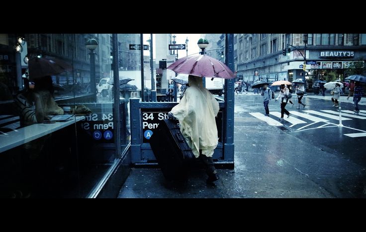 the image depicts a rainy day scene in what appears to be new york city.  the focus is on a person wearing a yellow raincoat and carrying a large suitcase, standing near a subway entrance at 34th street penn station.  numerous other pedestrians with umbrellas navigate the wet street crossing. the overall mood is dark and moody, with reflections visible in the glass of the nearby building and the wet pavement reflecting the city lights. the scene is full of atmospheric detail showcasing the daily life in a bustling urban setting during a downpour.