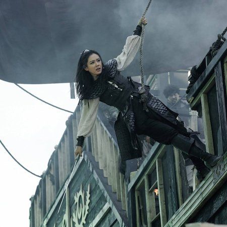 a woman dressed as a pirate is swinging on a rope while holding a sword. she is on a ship and appears to be in an action or adventure scene. the scene suggests danger and she looks like a warrior or a character in a movie or fantasy setting.