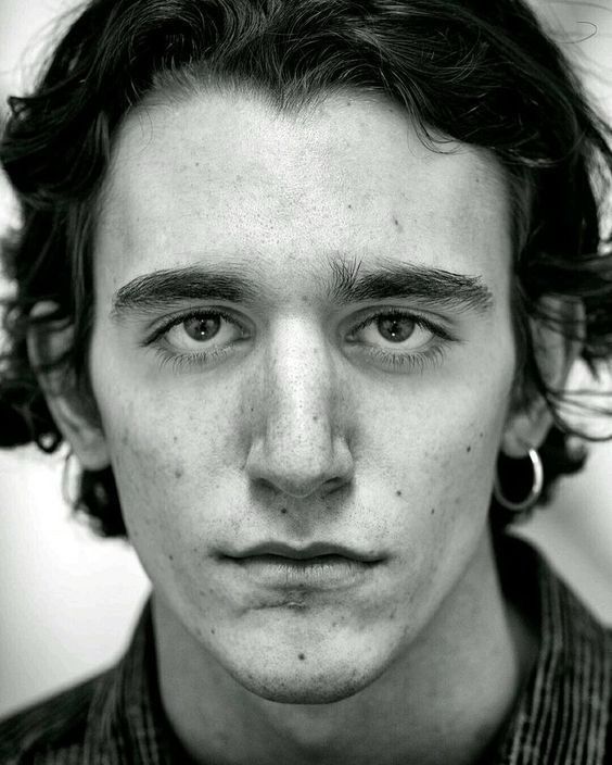 a black and white closeup portrait of a young man with dark, wavy hair. he has a serious, neutral expression on his face. he is wearing a hoop earring in his right ear. the image is sharply focused on his face, showing detail in his skin texture and hair. the lighting creates subtle shadows that add depth to the portrait.