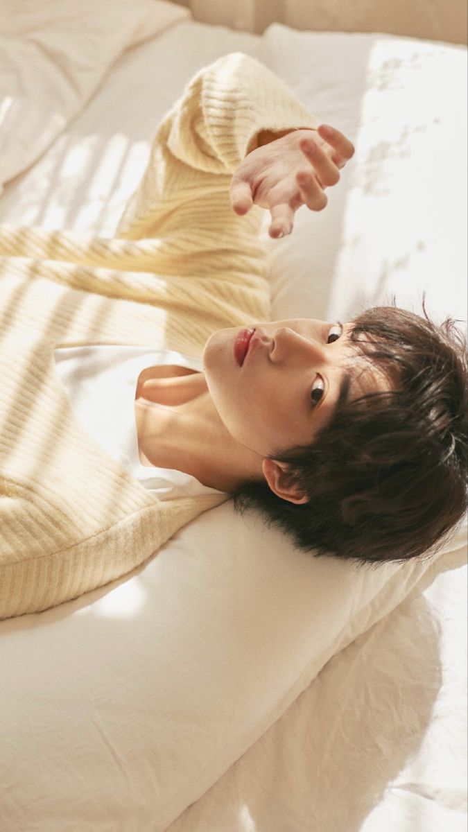 a young asian man with mediumlength dark hair is laying on a bed covered with a white sheet and a white comforter. he is wearing a beige colored knit cardigan and a white tshirt. the man is posing with his left arm reaching up towards the camera. the lighting in the image is soft and natural, coming from a window off to the right side of the frame, creating shadows of the window blinds across the man's body and the bed. the overall feeling of the image is one of relaxation and peace.