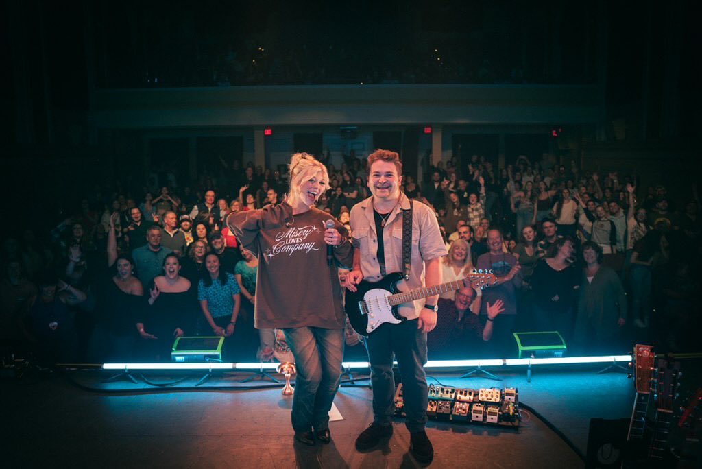 a photo captures a live music performance with a male musician holding a guitar and a female singer with a microphone on stage in front of a cheering crowd. the stage is lit with blue lights and various guitar effects pedals. the audience fills the theater, with many people smiling and raising their hands in excitement.