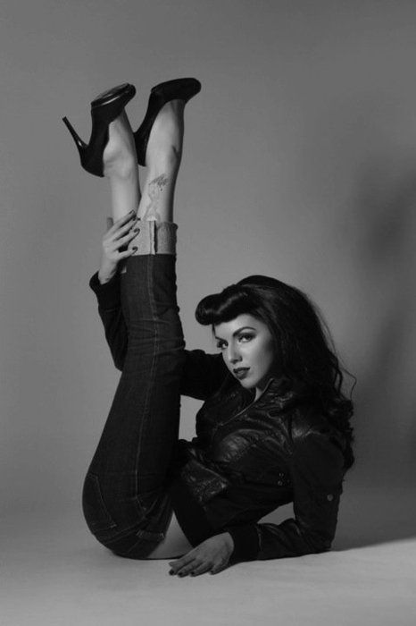 a woman in a black leather jacket and jeans strikes a retro pinup pose.  she has a classic pinup hairstyle and is wearing black heels.