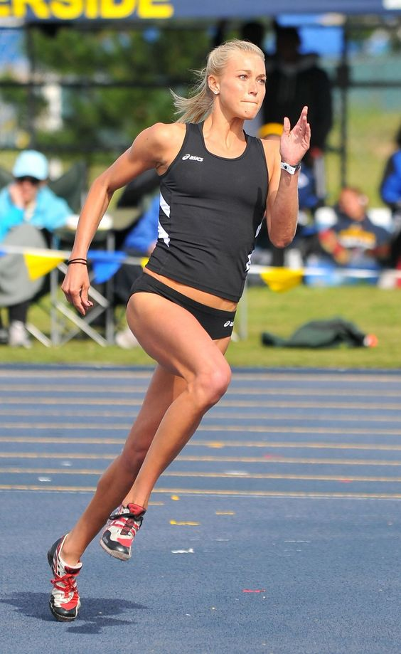 a female athlete in midstride while running a race on a blue track. she is wearing a black and white racing uniform and running shoes. the background is blurred and shows other people and the stadium setting.