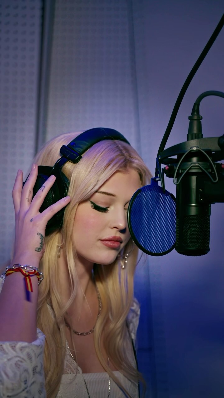 a young blonde woman is in a recording studio with headphones on and her hand adjusting them as she prepares to sing into a microphone with a pop filter. she is wearing jewelry and has a tattoo on her hand. the setting suggests she is a singer or performer involved in music production.