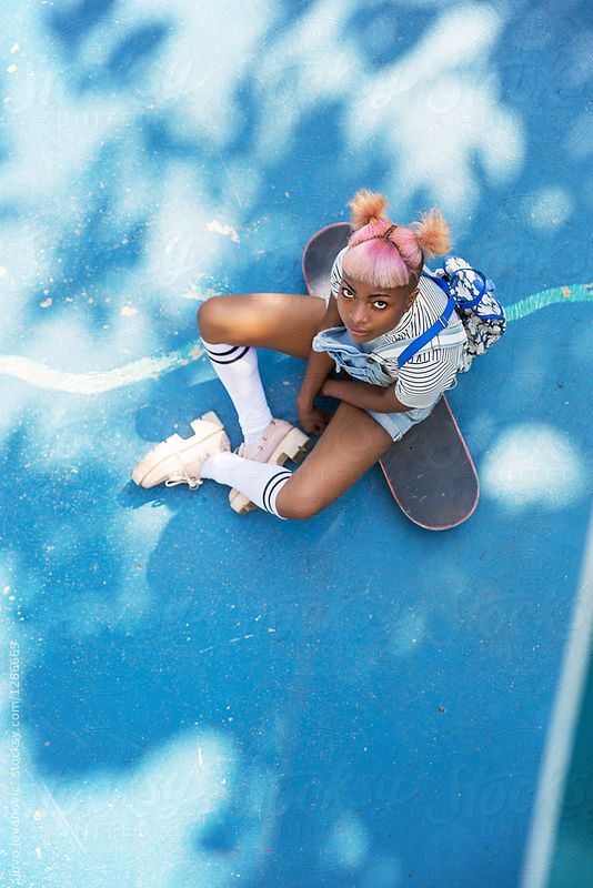 a teenage girl with pink hair sits on a skateboard and stares at the camera. she is wearing white socks with black stripes and white shoes with black accents, a striped shirt and blue denim overalls. the image is taken from a topdown angle, showcasing the blue background and the girl's pose.