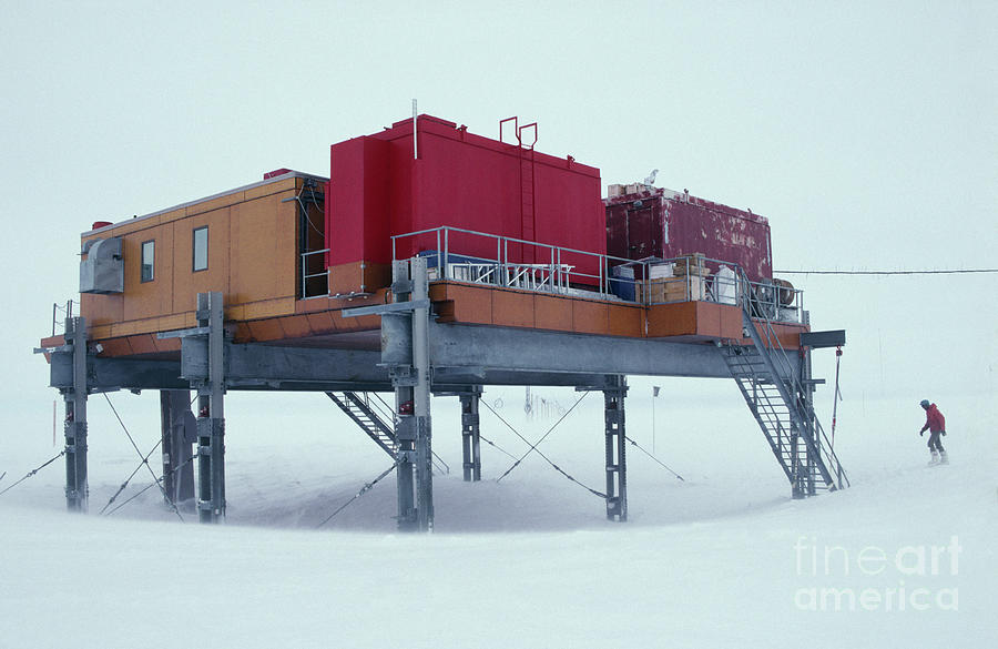 a red and yellow building, elevated on metal poles, is seen in a snowy landscape. it is an arctic research station, with cables and equipment for research. a single person in red clothing is walking towards the right side of the image, disappearing into the blizzard. the air is swirling with snow, making the building and landscape appear hazy.