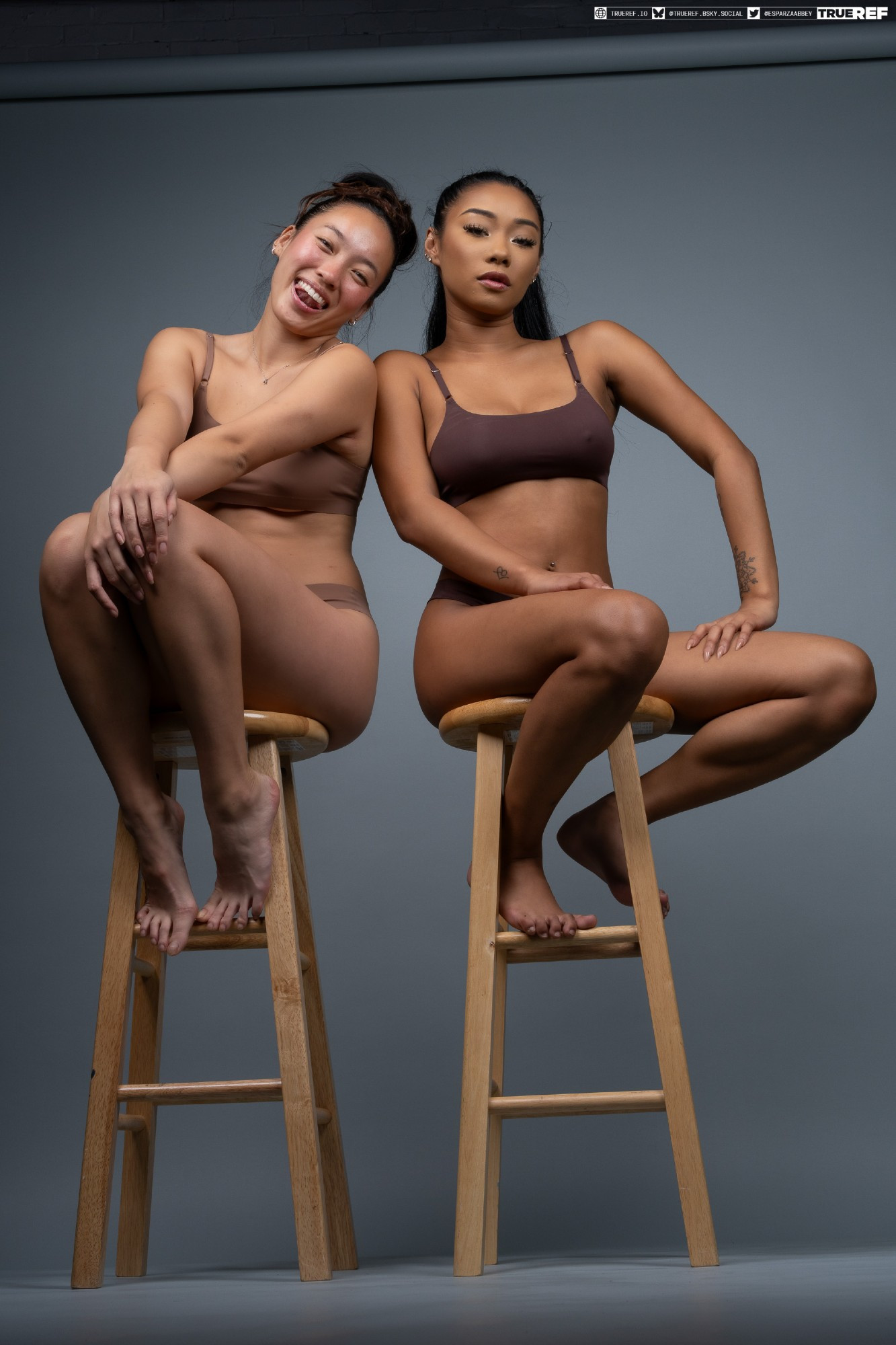 two asian women, one smiling and one with a more serious expression, sit confidently on wooden stools against a gray background. they are both wearing brown lingerie. the image is a studio shot showcasing their bodies in a way that celebrates body positivity and selflove.  the setting is simple, allowing the focus to remain on the women and their expressions. the overall feel is natural and relaxed.