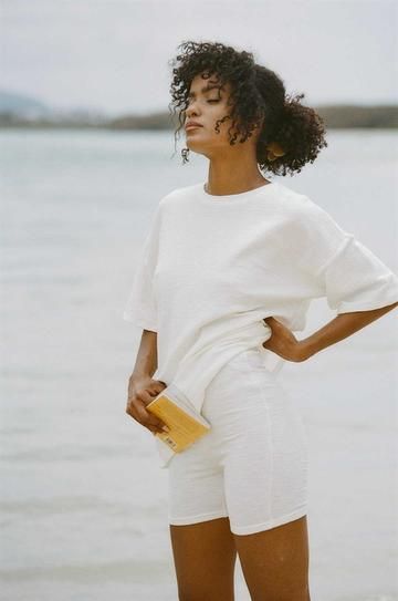 a woman with curly brown hair is standing on a beach wearing a white oversized tshirt and matching white shorts. she is holding a yellow book in her left hand and looking away from the camera. the photo is taken on a sunny day.