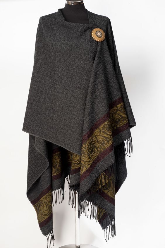 a gray shawl or cloak with a pattern near the edges and fringes at the bottom is displayed on a mannequin. the shawl is secured at the front with a large, circular brooch.