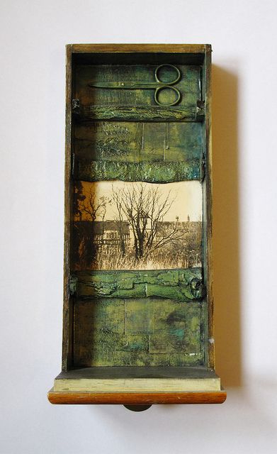 a mixed media assemblage artwork contained within a wooden drawerlike box. the box has a rustic and slightly distressed appearance, with a green textural background. inside the box, there is a pair of metal scissors at the top, a black and white photograph of a tree and building in the center, and more of the green textural background filling the spaces above and below the photo. the drawer box sits against a white wall, creating a shadow on the right side. the overall composition is a still life, combining found objects and artistic techniques.